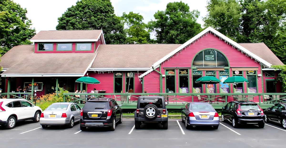 Nanola Restaurant in Malta. Red building. Outdoor deck with green umbrellas. Cars parked in parking lot.